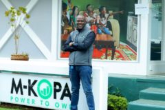 Babajide Duroshola standing in front of M-KOPA's logo