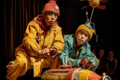 An AI-generated photo of two young men wearing raincoats on stage and playing with a toy. Created by the_real_nilspetter (@Nils P) via Midjourney.