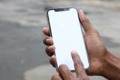 A person, Black or of African decent, holds a phone with a blank white screen in one hand and finger taps the screen with the other hand