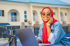 An African freelancer sitting in front of her laptop and smiling to the camera (the viewer) with tinted shades on.