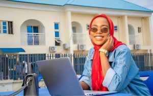 An African freelancer sitting in front of her laptop and smiling to the camera (the viewer) with tinted shades on.