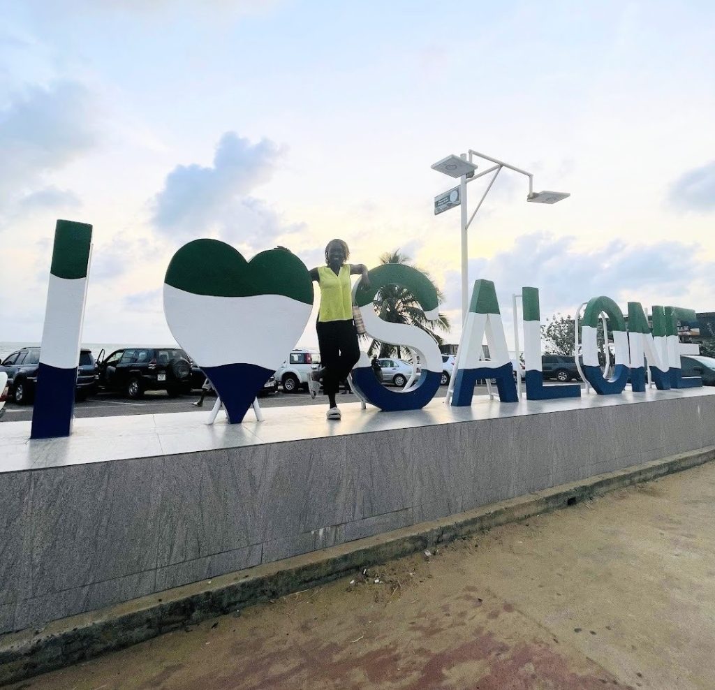 Oke poses for a photo at the ‘I Love Salone’ sign in Freetown, Sierra Leone.